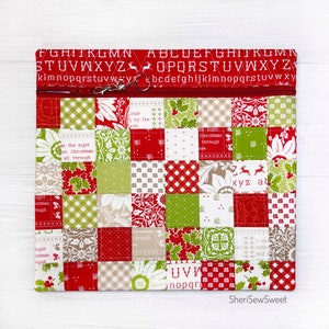 May include: A red and green patchwork fabric zipper pouch with a zipper pull and a white background. The fabric has a variety of Christmas-themed patterns, including snowflakes, houses, and reindeer. The pouch is lined with a red fabric and has a white zipper. The text "SheriSewSweet" is printed on the bottom of the pouch.