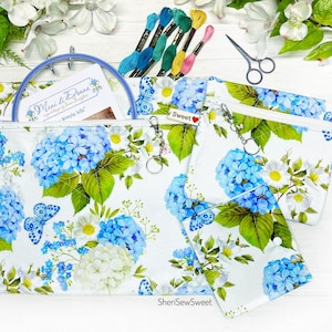 May include: A set of three zippered fabric pouches with a floral hydrangea pattern in blue, white, and green. The pouches are accompanied by embroidery floss, a hoop, and small scissors. The text "SheriSewSweet" is visible.