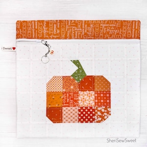 May include: A white zippered pouch with a patchwork pumpkin design. The pumpkin is made of orange, red, and green fabric squares. The pouch has a white background with small pink dots. The zipper has a silver pull with a small charm.