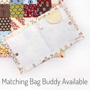 May include: Open, fabric-lined pouch with a white interior and a decorative patterned border. The pouch is secured with a snap closure and features a small, scalloped, beige felt piece. The background shows a patchwork quilt.