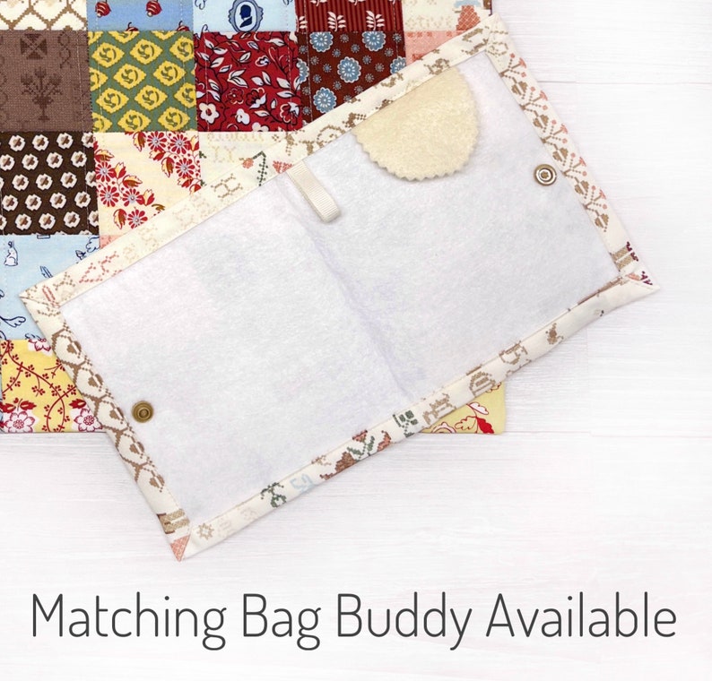 May include: Open, fabric-lined pouch with a white interior and a decorative patterned border. The pouch is secured with a snap closure and features a small, scalloped, beige felt piece. The background shows a patchwork quilt.