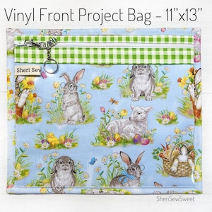 May include: A clear vinyl project bag with a blue and white fabric lining featuring a bunny, lamb, and chick pattern. The bag has a green and white checkered zippered pocket and measures 11 inches by 13 inches. The bag is labeled "Sheri Sew Sweet".