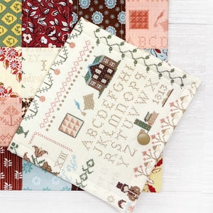 May include: A cream-colored, cross-stitch style fabric case with a button closure, featuring a detailed pattern of houses, alphabets, and decorative borders. The case is layered over a patchwork quilt with floral and geometric patterns in various colors.