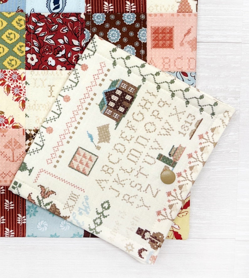 May include: A cream-colored, cross-stitch style fabric case with a button closure, featuring a detailed pattern of houses, alphabets, and decorative borders. The case is layered over a patchwork quilt with floral and geometric patterns in various colors.