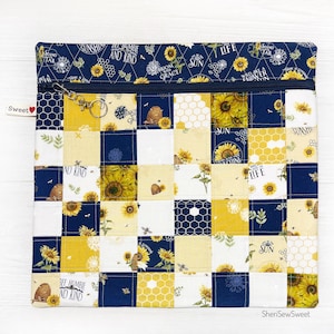 May include: A zippered pouch with a patchwork design featuring yellow sunflowers, honeycomb patterns, and blue and white floral prints. The pouch has a zipper closure and a small key chain attached to the zipper pull. The fabric has text printed on it, including "Be Humble and Kind", "Bloom with Grace", "Sun", "Life", "Stand Tall", "Wild like a Flower", and "Sunshine".