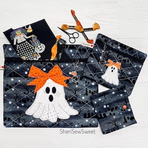 May include: Three zippered pouches in a black fabric with a white graveyard and ghost pattern. The largest pouch features a white ghost applique with an orange polka dot bow. Includes black embroidery scissors, orange thread, and a cross-stitch project.