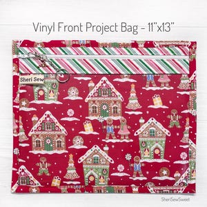 May include: A red vinyl project bag, 11" x 13", with a zipper closure. The bag features a festive gingerbread house pattern and a striped top border. The text "Vinyl Front Project Bag - 11"x13"" is printed on the bag.