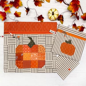 May include: Two zippered pouches with a patchwork pumpkin design. The larger pouch has a white and black striped background with a patchwork pumpkin in the center. The smaller pouch has a white and black striped background with a solid orange pumpkin in the center. Both pouches have orange zippers and a small orange tag that says "Sweet".