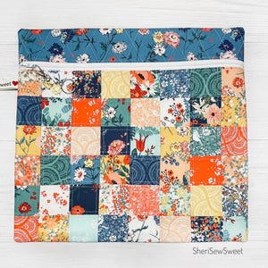 May include: A patchwork zippered pouch with a floral design. The pouch features a variety of colorful floral patterns in squares, with a blue floral top and a white zipper. The text "SheriSewSweet" is visible.