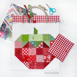 May include: A white fabric pouch with a patchwork apple design in red and green squares. The pouch is open, revealing a red and white gingham lining, embroidery floss, and a wooden embroidery hoop. A small gingham square and scissors are also visible.