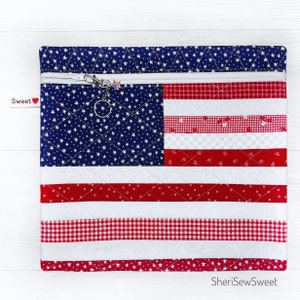 May include: A quilted zippered pouch featuring a design resembling the American flag. The pouch has a blue field with white stars and red and white stripes. A silver zipper and a small charm are visible. The text "Sweet" is on a tag.