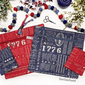 May include: Red and navy blue zippered pouches and small square items with patriotic themes. The pouches feature the number "1776" and text. The small items have star and text designs. A pair of scissors and embroidery floss are also visible.