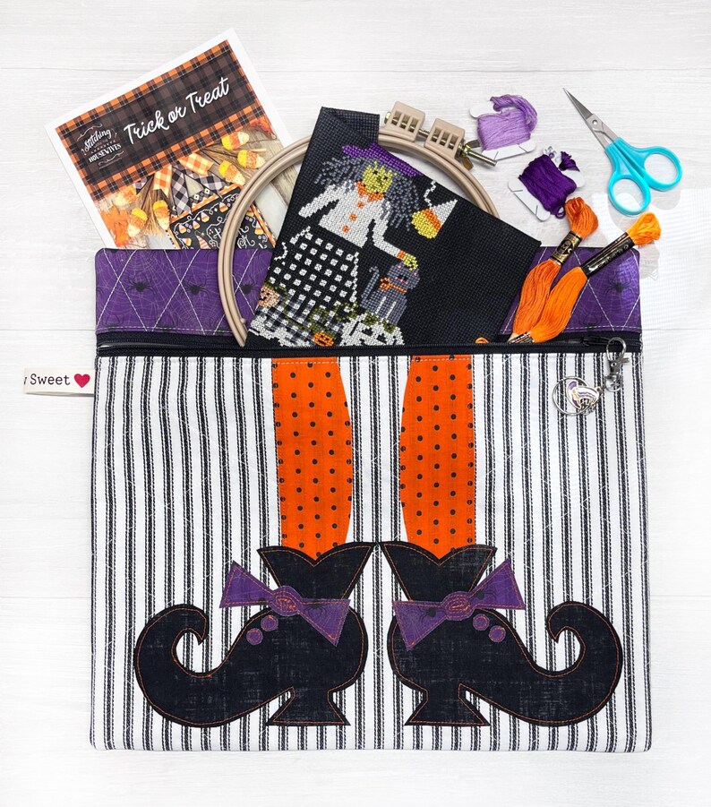 May include: A black and white striped zippered pouch with a witch's legs and shoes design. The pouch is filled with a cross-stitch project, embroidery floss, and scissors. The pouch has a purple heart tag with the text "Sweet" on it.
