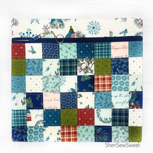 May include: A quilted pouch with a patchwork design featuring winter-themed patterns in shades of blue, green, and red. The top section has a white background with snowflakes and a zipper closure. A snowflake charm hangs from the zipper.