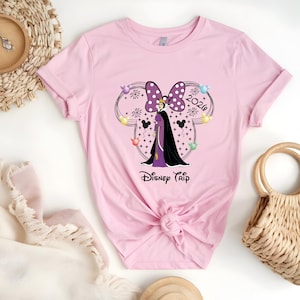 May include: A light pink t-shirt featuring a graphic of a Disney character with a purple bow, the text "2020", and the words "Disney Trip". The design includes black Mickey Mouse silhouettes and colorful balloons.