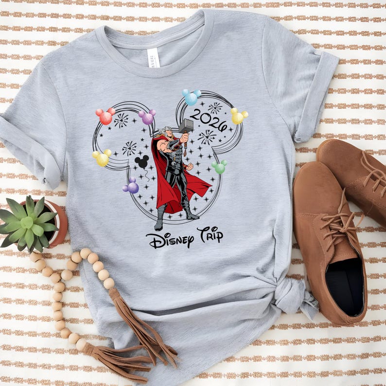 May include: A light gray t-shirt featuring a graphic of Thor holding his hammer inside a Mickey Mouse head outline. The shirt has the text "2026" and "Disney Trip" in black. Colorful balloons and fireworks complete the design.