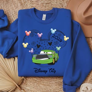 May include: Royal blue sweatshirt featuring a green cartoon race car with the text "Disney Trip". The design includes Mickey Mouse ears, colorful balloons, fireworks, and the year "2026".
