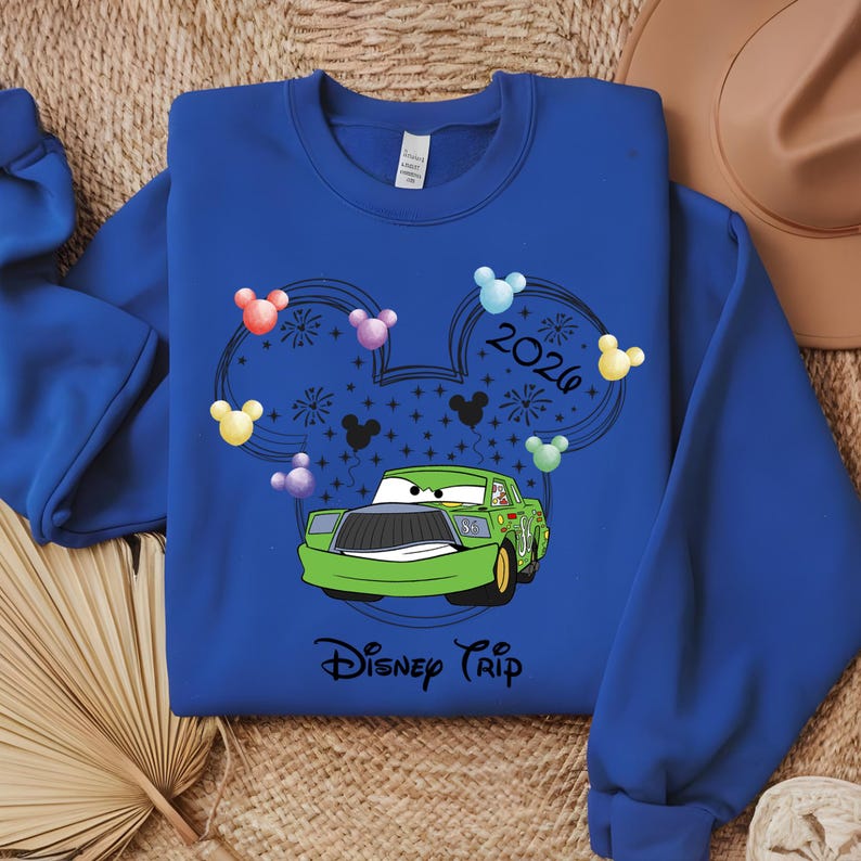 May include: Royal blue sweatshirt featuring a green cartoon race car with the text "Disney Trip". The design includes Mickey Mouse ears, colorful balloons, fireworks, and the year "2026".
