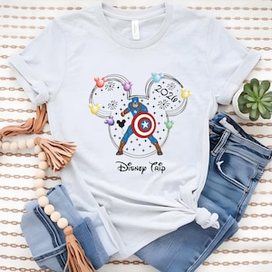 May include: White t-shirt featuring a Captain America graphic within a Mickey Mouse head outline, with the text "2026" and "Disney Trip". The design includes colorful balloons and fireworks. The shirt is paired with blue jeans.