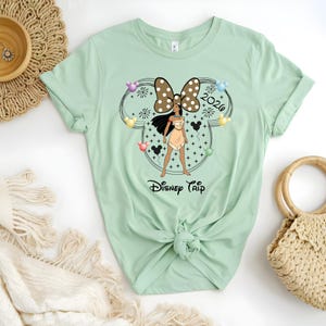 May include: A light green t-shirt featuring a graphic of a character in a Minnie Mouse ear design with a brown polka dot bow, the text "2020", and the words "Disney Trip". The character is wearing a tan dress.
