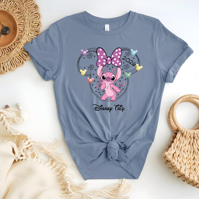 May include: A blue t-shirt featuring a pink cartoon character with a polka-dot bow, surrounded by a Mickey Mouse-shaped design with the text "2026" and "Disney Trip". The design includes colorful accents and stars.