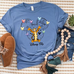 May include: A light blue t-shirt featuring a graphic of Tigger from Winnie the Pooh inside a Mickey Mouse head outline. The design includes the text "2020" and "Disney Trip". Colorful Mickey Mouse head balloons and fireworks are also included.