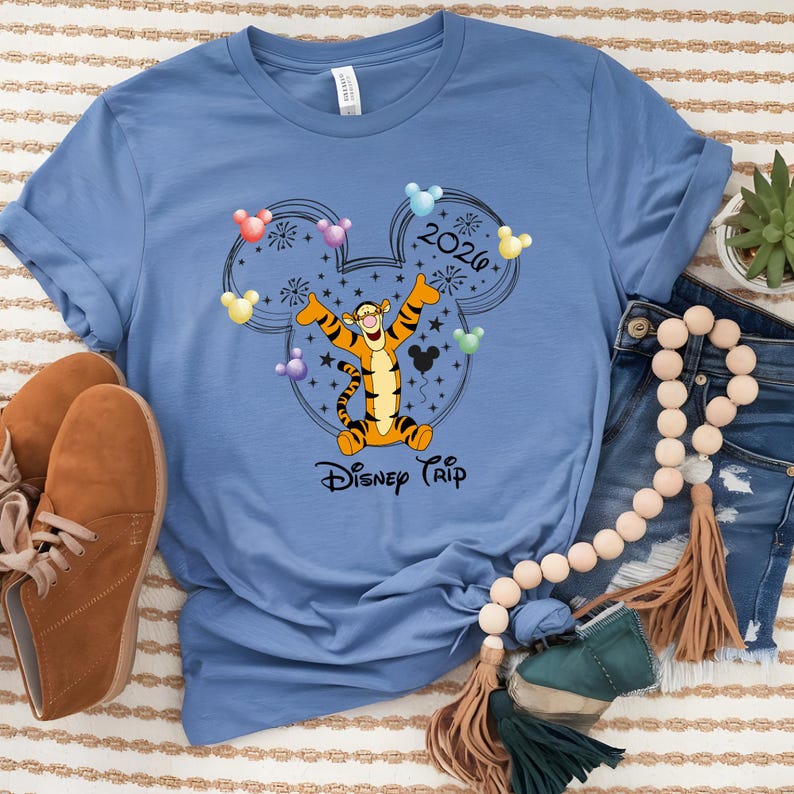 May include: A light blue t-shirt featuring a graphic of Tigger from Winnie the Pooh inside a Mickey Mouse head outline. The design includes the text "2020" and "Disney Trip". Colorful Mickey Mouse head balloons and fireworks are also included.