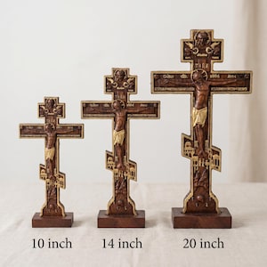 Puede incluir: Tres cruces ortodoxas de madera de diferentes tamaños: 25, 35 y 51 cm de altura. Cada cruz presenta una figura tallada de Jesucristo con detalles dorados, sobre madera marrón oscura. Las cruces están sobre una base de madera.