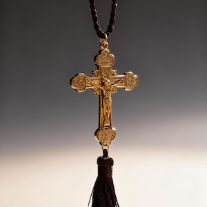 May include: A gold-coloured crucifix pendant with a detailed depiction of Jesus Christ on the cross. The cross is suspended from a brown cord necklace and has a matching tassel at the bottom. The background is a gradient of grey and white.