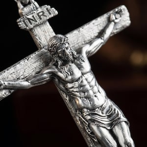 Könnte beinhalten: Ein silberfarbenes Metallkruzifix mit einer detaillierten Jesusfigur auf einem strukturierten Kreuz. Am oberen Teil des Kreuzes befindet sich eine kleine Plakette mit der Inschrift INRI. Das Metall hat ein antikes Finish.