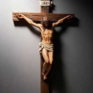 Può includere: Scultura dettagliata di Gesù Cristo su una croce di legno. La figura è raffigurata con una corona di spine, un perizoma e ferite visibili. La croce ha uno stendardo con l'iscrizione "INRI". La scultura è ambientata su uno sfondo grigio.