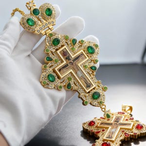 Puede incluir: Colgante de cruz religiosa dorada adornado con piedras preciosas verdes esmeralda y rojas. La cruz presenta una imagen central de Jesucristo y está sostenida por una mano enguantada. Una cruz más pequeña es visible debajo.
