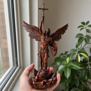Op de afbeelding: Gedetailleerd bruin beeld van een engel met grote vleugels, die een staf met kruis en een schild vasthoudt. De engel staat boven een figuur. Het beeld is ongeveer 25 cm hoog.