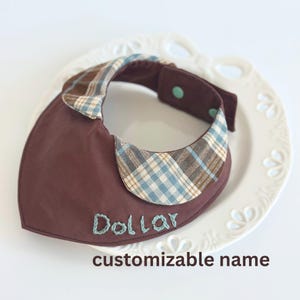Puede incluir: Un babero marrón con un cuello a cuadros en tonos azules, marrones y blancos. La palabra "Dollar" está bordada con hilo azul claro. El texto "customizable name" está debajo.
