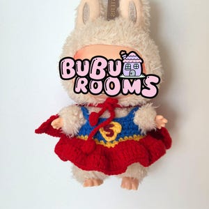 May include: A plush doll wearing a superhero costume. The doll has a fluffy, cream-colored bunny hat and a red, blue, and yellow crocheted outfit. The text "BUBU ROOMS" is across the doll's face.