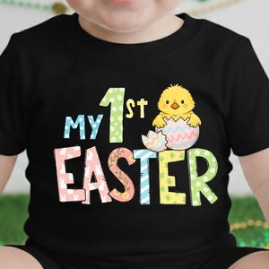 May include: Black t-shirt with the text "My 1st Easter" in colorful letters and a graphic of a yellow chick hatching from an egg. The shirt is designed for a baby or toddler.