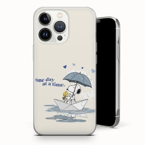 以下が含まれることがあります： 傘の下の紙のボートに乗ったスヌーピーとウッドストックの漫画イラストが描かれた携帯電話ケース。「one day at a time」の文字と青いハートのアクセントが付いています。