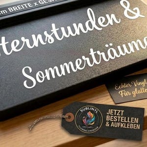 Könnte beinhalten: Ein schwarzes rechteckiges Schild mit weißem Schreibschrifttext Sternstunden & Sommerträume und einem 25 cm Breitenetikett. Ein schwarzer Anhänger mit buntem Federlogo und dem Text JETZT BESTELLEN & AUFKLEBEN ist mit einer Schnur befestigt.