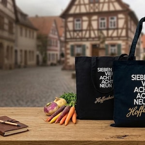 Könnte beinhalten: Drei schwarze Stofftaschen mit weißem Aufdruck SIEBEN VIER ACHT ACHT NEUN und goldener Schreibschrift mit der Aufschrift Hoffenheim oder Obrigheim, präsentiert neben einem kleinen braunen Notizbuch mit Stift und einem Bündel frischem Gemüse.