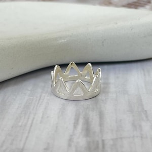 Può includere: Un anello color argento con un design a corona. L'anello presenta una serie di ritagli triangolari che formano la forma della corona. L'anello è esposto su una superficie chiara, mettendo in risalto la sua lucentezza metallica.