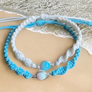 May include: Two macrame bracelets with opal-like stones. One bracelet is white, the other is light blue. Both have a silver wire setting around the stones. The bracelets are on a light tan surface with a wave-like background.