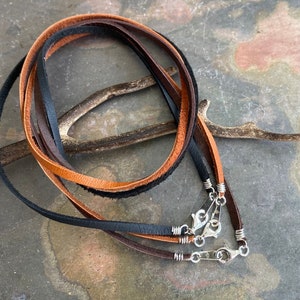 May include: Three leather cords in brown, black, and tan with silver clasps. The cords are arranged in a circle and are lying on a textured surface.