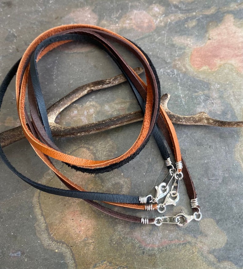 May include: Three leather cords in brown, black, and tan with silver clasps. The cords are arranged in a circle and are lying on a textured surface.
