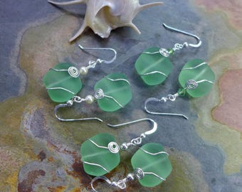 May include: Sea glass earrings with a light green hue, wire-wrapped in a floral design. The earrings have a silver hook and are paired with a seashell. The earrings are a unique accessory.