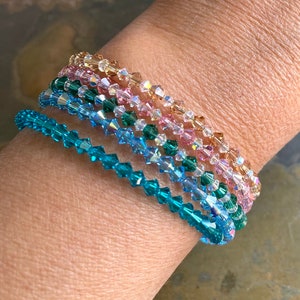 May include: A set of five crystal bracelets in shades of teal, blue, pink, and gold. The bracelets are stacked on a person's wrist.