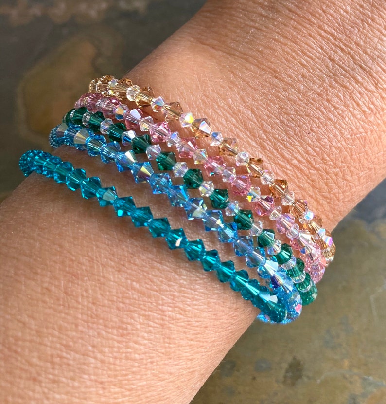 May include: A set of five crystal bracelets in shades of teal, blue, pink, and gold. The bracelets are stacked on a person's wrist.