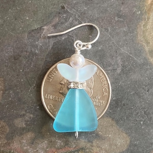 May include: A handcrafted earring featuring a sea glass angel design. The angel has a light blue body, a white halo, and a pearl head. The earring is attached to a silver hook. The angel is approximately 1 inch in height.