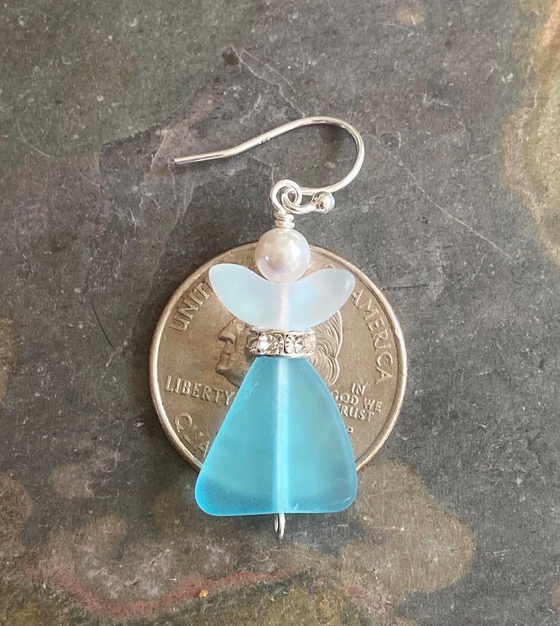 May include: A handcrafted earring featuring a sea glass angel design. The angel has a light blue body, a white halo, and a pearl head. The earring is attached to a silver hook. The angel is approximately 1 inch in height.