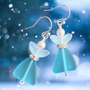 May include: A pair of angel-shaped earrings with a light blue body and wings. The earrings have silver-colored hooks and are adorned with a pearl and a band of clear rhinestones. The background is a blurred blue with white bokeh.