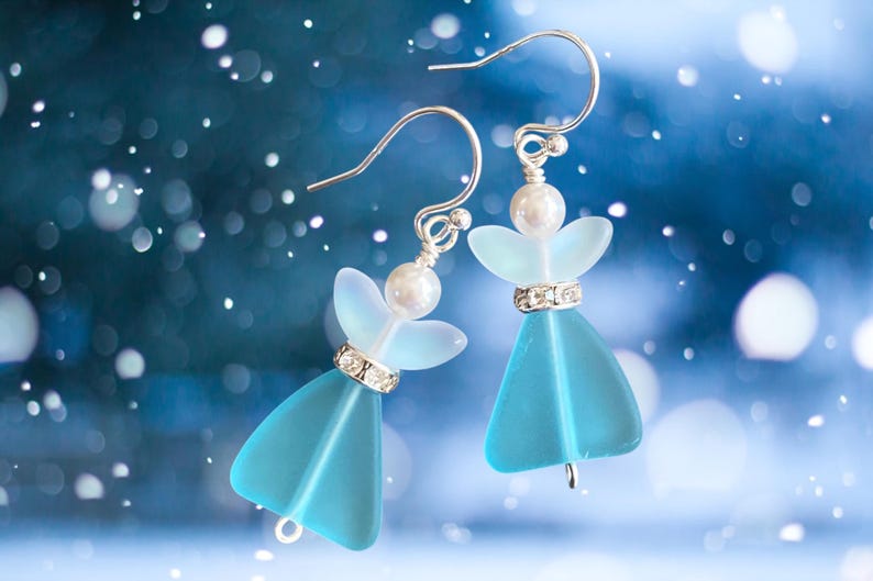 May include: A pair of angel-shaped earrings with a light blue body and wings. The earrings have silver-colored hooks and are adorned with a pearl and a band of clear rhinestones. The background is a blurred blue with white bokeh.